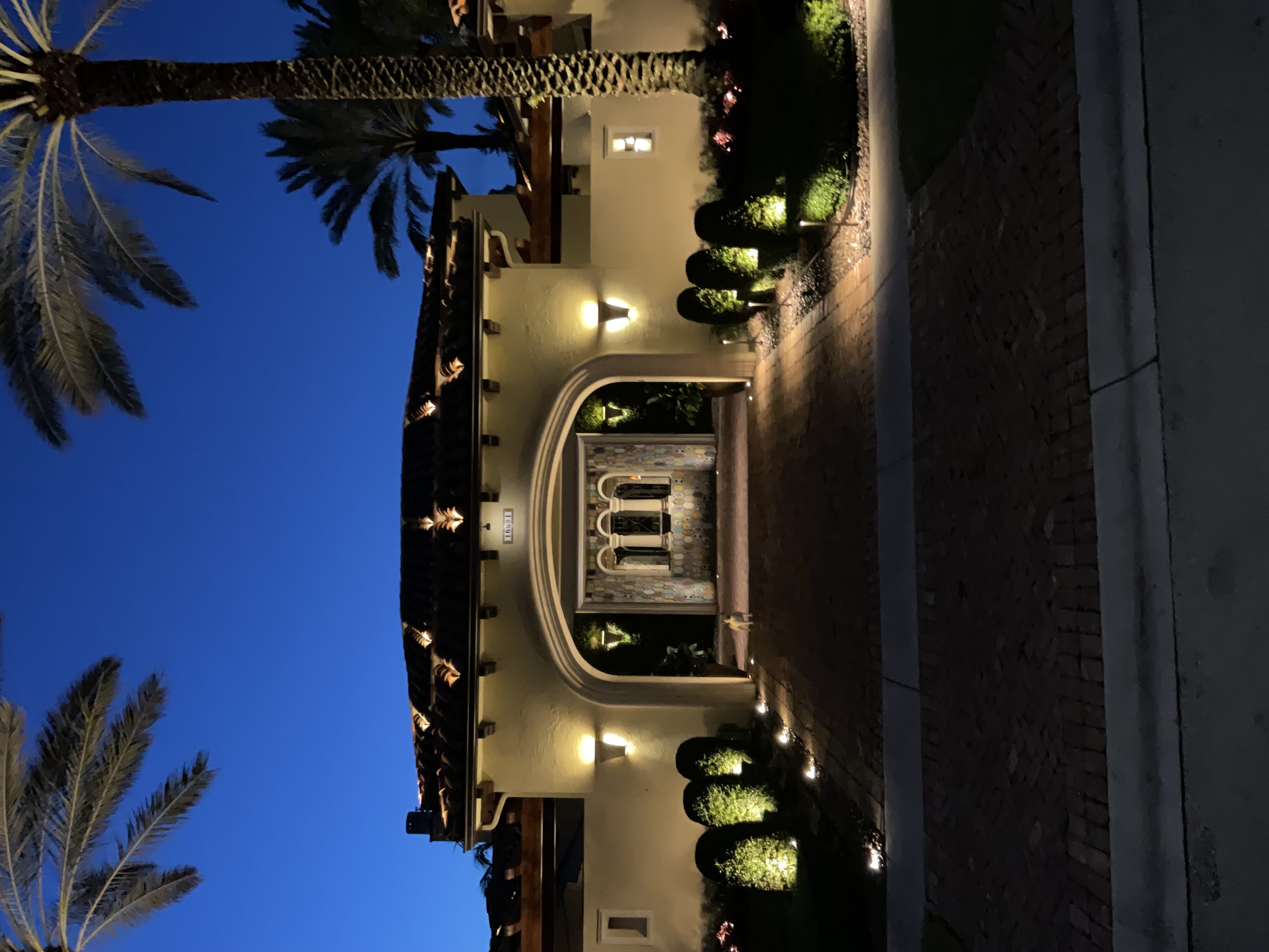 Nighttime entry sequence of the residence framed by palms, landscape lighting, and an arched gatehouse