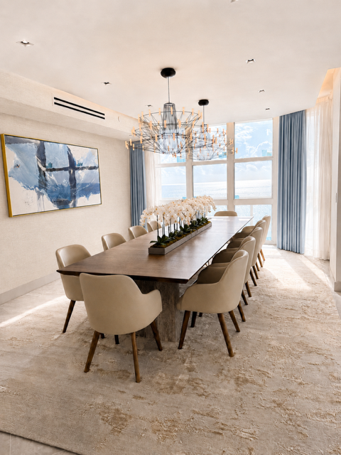 Oceanfront dining room with long wood table, upholstered chairs, chandelier, blue drapery, art, and water views